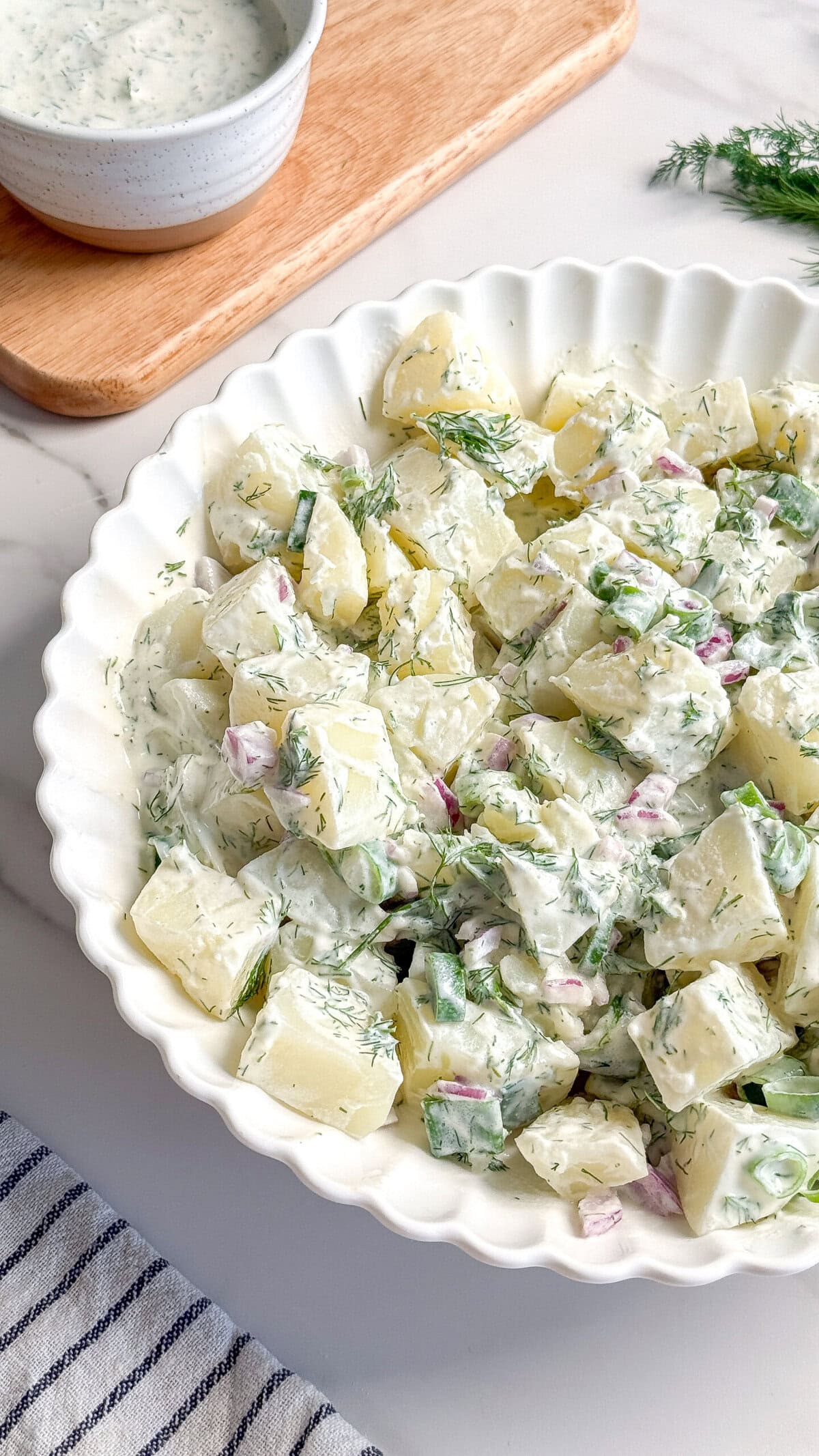 A white scalloped bowl filled with creamy Vegan Potato Salad garnished with fresh herbs and diced red onions, with a bowl of Vegan Tzatziki and sprigs of dill on a wooden board in the background.