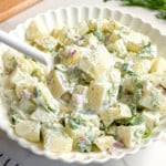 A white scalloped bowl filled with creamy Vegan Potato Salad, garnished with fresh dill and red onion, sits on a white surface. A serving spoon is in the bowl. A striped towel and a small bowl of Vegan Tzatziki are nearby.