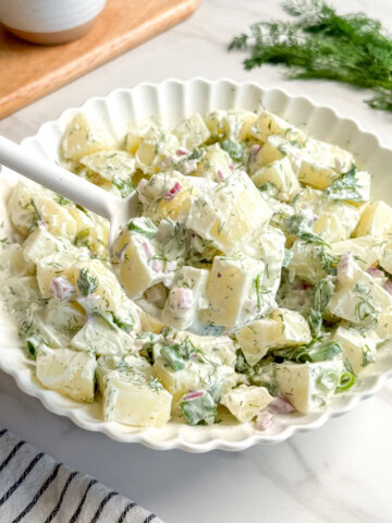 A white scalloped bowl filled with creamy Vegan Potato Salad, garnished with fresh dill and red onion, sits on a white surface. A serving spoon is in the bowl. A striped towel and a small bowl of Vegan Tzatziki are nearby.