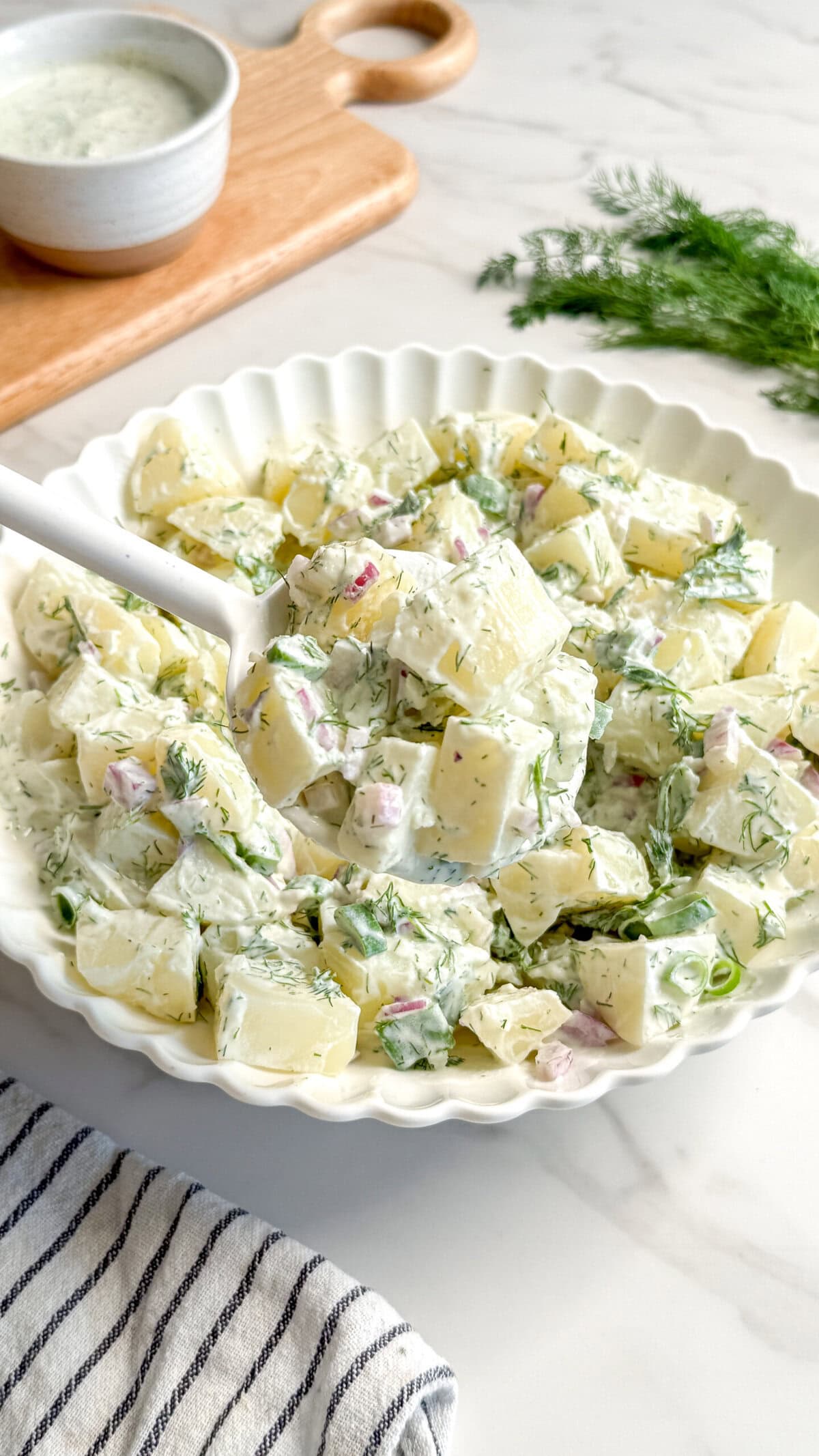 A white scalloped bowl filled with creamy Vegan Potato Salad, garnished with fresh dill and red onion, sits on a white surface. A serving spoon is in the bowl. A striped towel and a small bowl of Vegan Tzatziki are nearby.