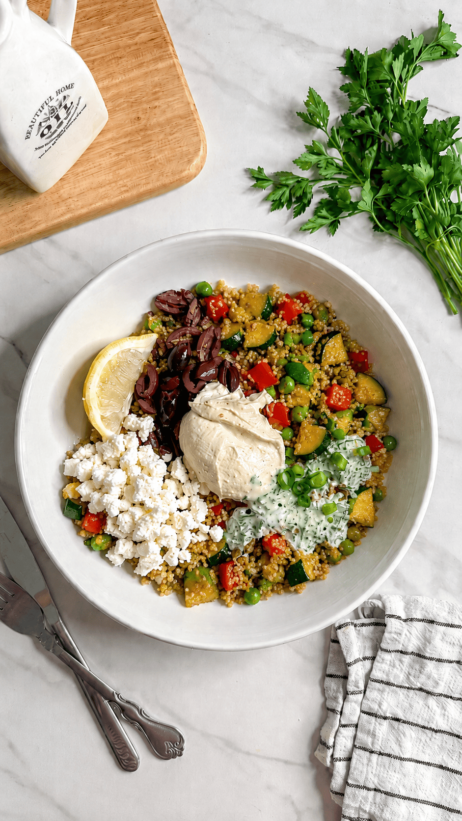 A Vegan Mediterranean Quinoa Bowl featuring hummus, feta cheese, chopped vegetables, olives, tzatziki sauce, and a lemon wedge. Parsley, a striped napkin, cutlery, and a cutting board rest nearby on a white surface.