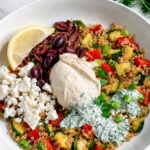A Vegan Mediterranean Quinoa Bowl features a white bowl filled with quinoa, diced zucchini, red bell pepper, kalamata olives, hummus, fresh parsley, and more-perfectly topped with a lemon wedge for a burst of flavor.