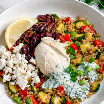 A Vegan Mediterranean Quinoa Bowl features a white bowl filled with quinoa, diced zucchini, red bell pepper, kalamata olives, hummus, fresh parsley, and more—perfectly topped with a lemon wedge for a burst of flavor.