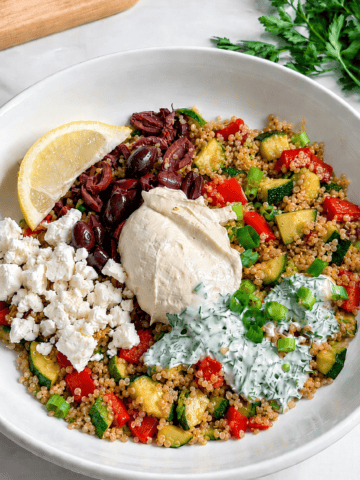 A Vegan Mediterranean Quinoa Bowl features a white bowl filled with quinoa, diced zucchini, red bell pepper, kalamata olives, hummus, fresh parsley, and more-perfectly topped with a lemon wedge for a burst of flavor.