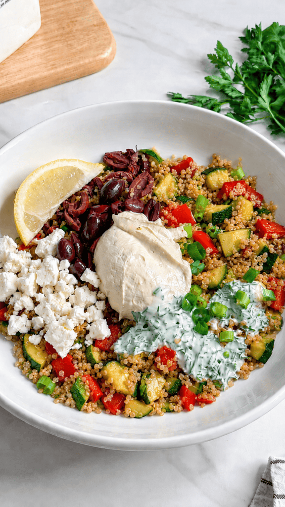 A Vegan Mediterranean Quinoa Bowl features a white bowl filled with quinoa, diced zucchini, red bell pepper, kalamata olives, hummus, fresh parsley, and more—perfectly topped with a lemon wedge for a burst of flavor.
