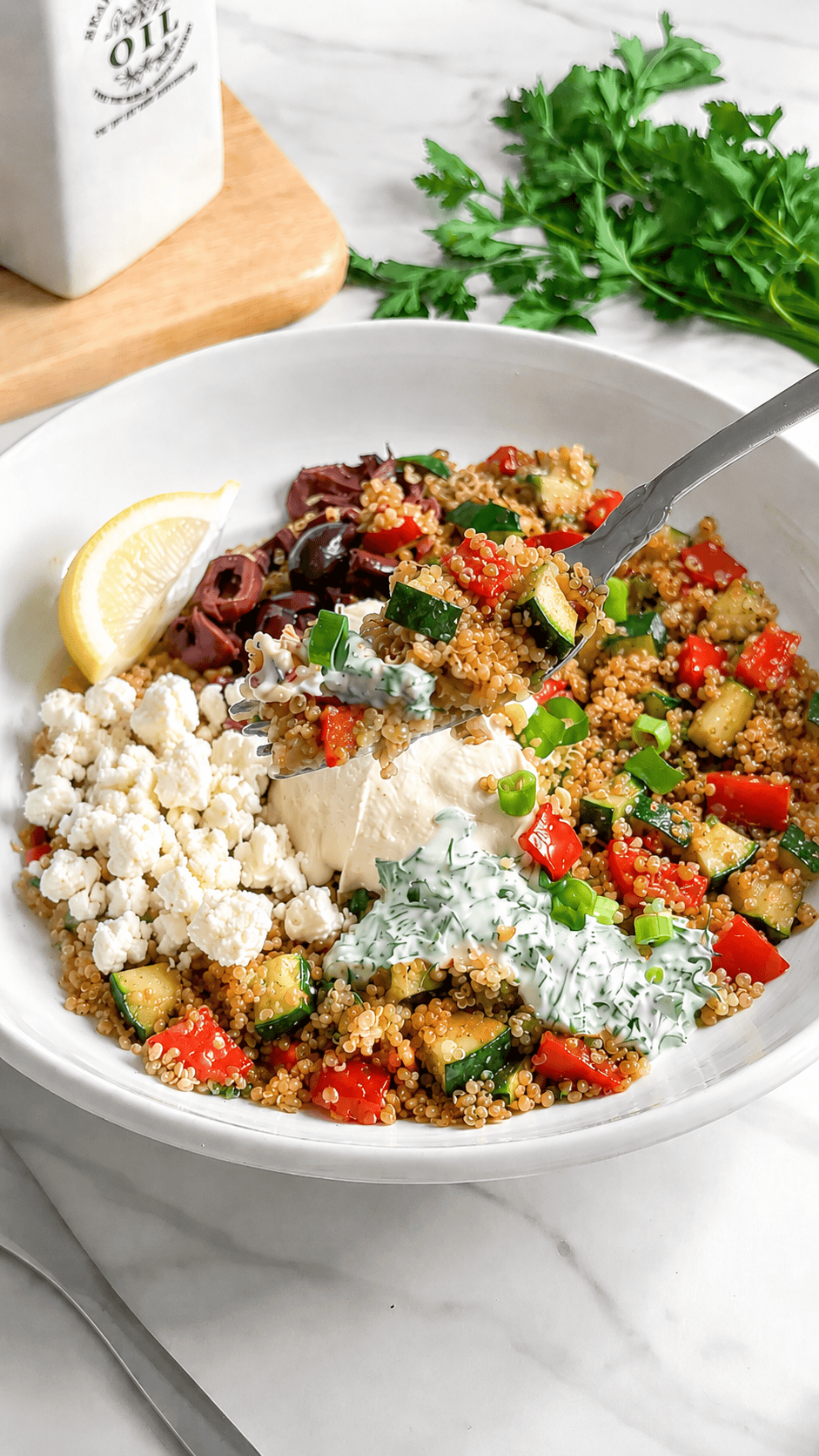 A Vegan Mediterranean Quinoa Bowl with chopped zucchini, red bell pepper, olives, hummus, and green onions, garnished with a lemon wedge and served in a white bowl on a marble surface.