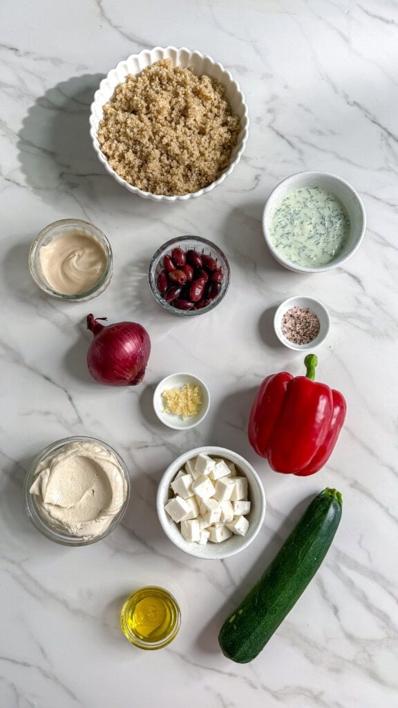 Various fresh and prepared ingredients for a Vegan Mediterranean Quinoa Bowl are arranged on a white marble surface, including quinoa, kidney beans, feta cheese, red onion, red bell pepper, zucchini, olive oil, garlic, sauces, and spices in bowls and jars.