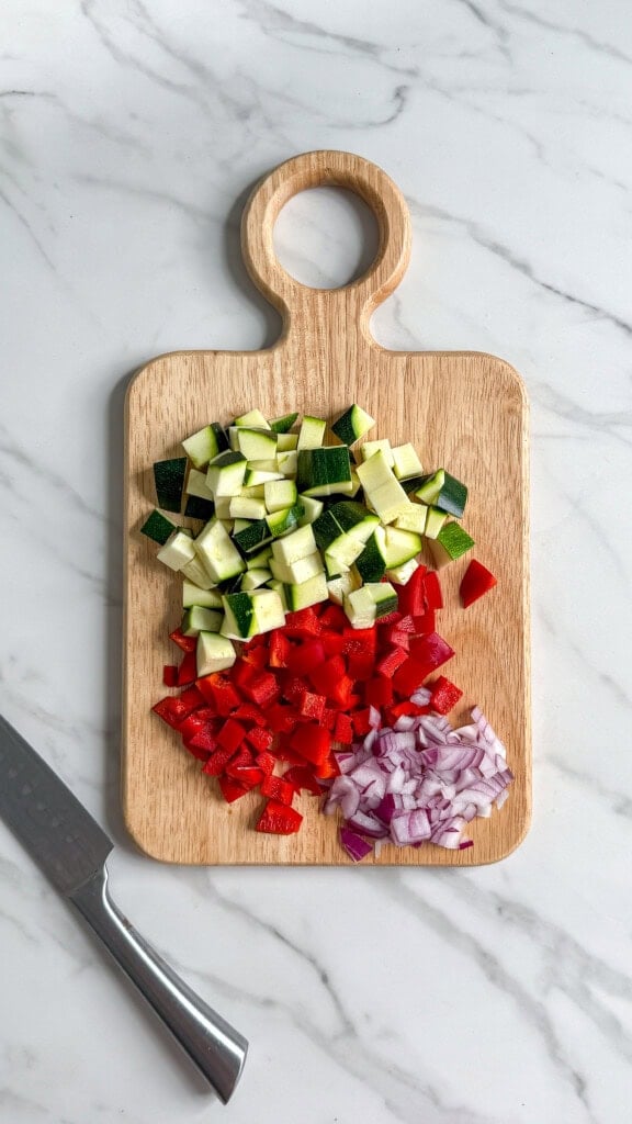 A wooden cutting board with diced zucchini, red bell pepper, and red onion, arranged in sections-perfect veggies for a Vegan Mediterranean Quinoa Bowl. A kitchen knife lies to the left on a white marble surface.