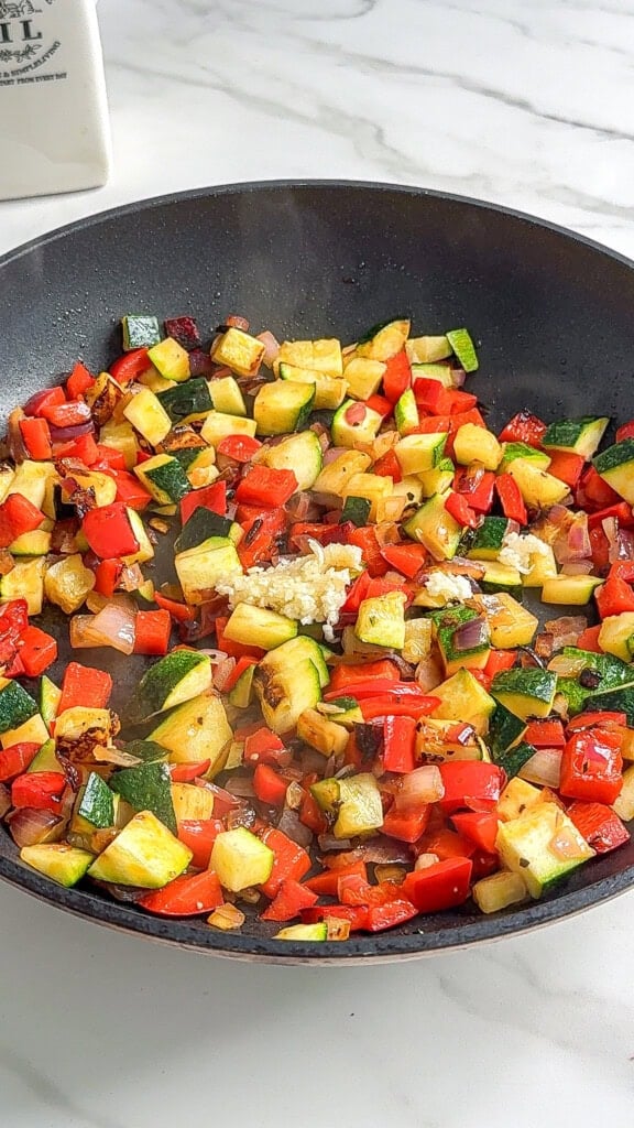 A black frying pan on a white countertop filled with sautéed diced red bell pepper, zucchini, onions, and minced garlic-perfect veggies for a Vegan Mediterranean Quinoa Bowl. The vegetables are lightly browned and steaming.