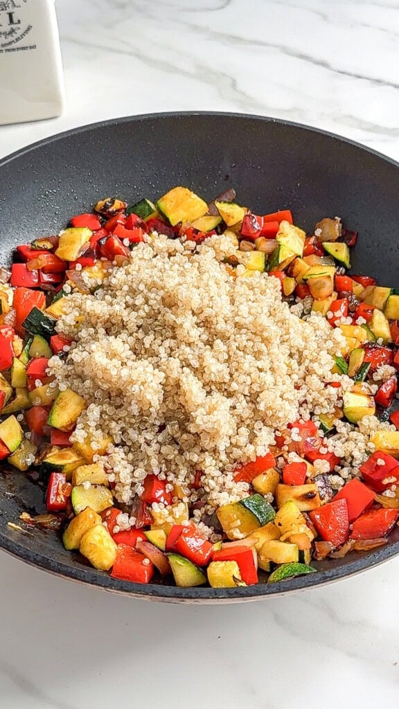 A Vegan Mediterranean Quinoa Bowl featuring sautéed diced zucchini, red bell peppers, and onions in a skillet, topped with a mound of cooked quinoa on a white marble countertop.