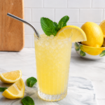 A glass of Easy Fresh Squeezed Refined Sugar-Free Lemonade with crushed ice, a metal straw, mint leaves, and a lemon slice sits on a marble board next to lemon wedges and mint leaves. Whole lemons are in a bowl in the background.