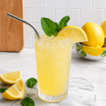 A glass of Easy Fresh Squeezed Refined Sugar-Free Lemonade with crushed ice, a metal straw, mint leaves, and a lemon slice sits on a marble board next to lemon wedges and mint leaves. Whole lemons are in a bowl in the background.