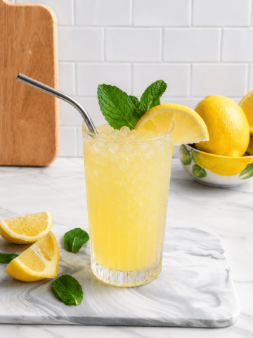 A glass of Easy Fresh Squeezed Refined Sugar-Free Lemonade with crushed ice, a metal straw, mint leaves, and a lemon slice sits on a marble board next to lemon wedges and mint leaves. Whole lemons are in a bowl in the background.
