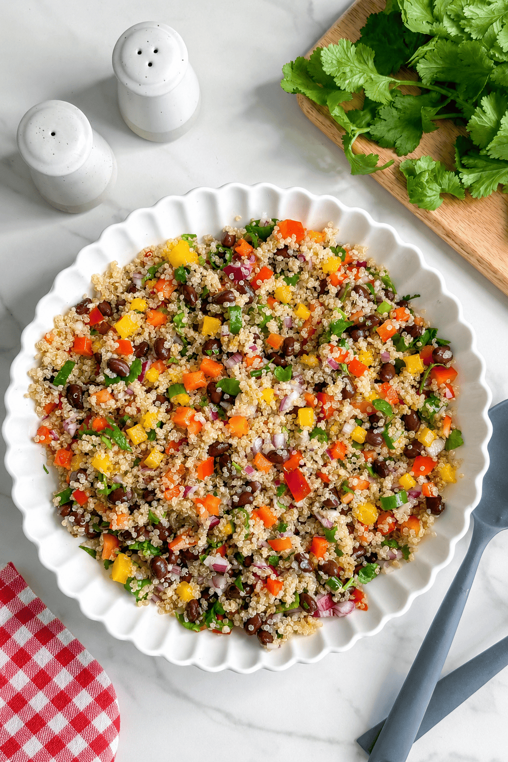 A white dish filled with vibrant Mexican Quinoa Salad featuring black beans, red onion, bell peppers, and herbs, sits on a marble surface beside salt and pepper shakers, cilantro, utensils, and a red-checkered napkin.