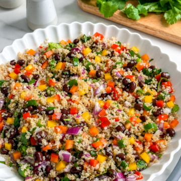 A white scalloped bowl filled with vibrant Mexican Quinoa Salad featuring black beans, chopped bell peppers, red onion, and herbs. In the background, fresh cilantro and salt and pepper shakers sit on a marble counter.