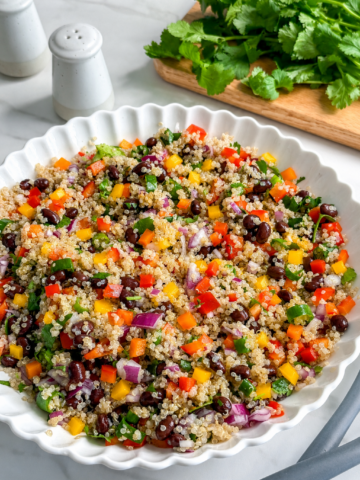 A white scalloped bowl filled with vibrant Mexican Quinoa Salad featuring black beans, chopped bell peppers, red onion, and herbs. In the background, fresh cilantro and salt and pepper shakers sit on a marble counter.