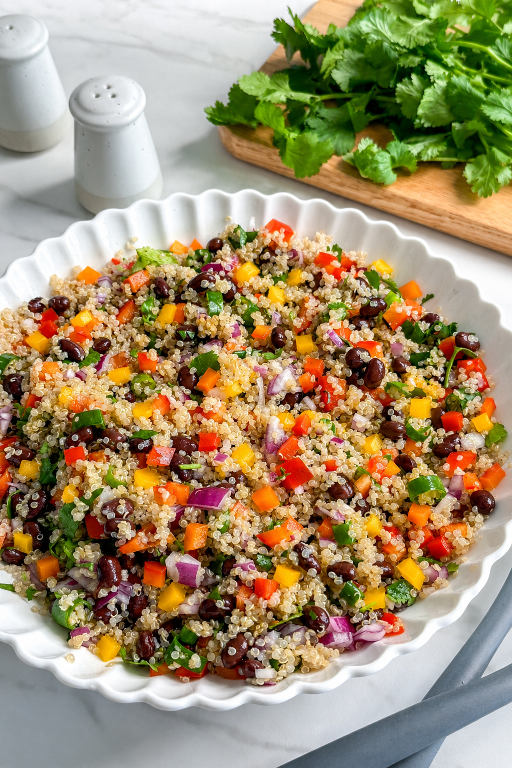 A white scalloped bowl filled with vibrant Mexican Quinoa Salad featuring black beans, chopped bell peppers, red onion, and herbs. In the background, fresh cilantro and salt and pepper shakers sit on a marble counter.