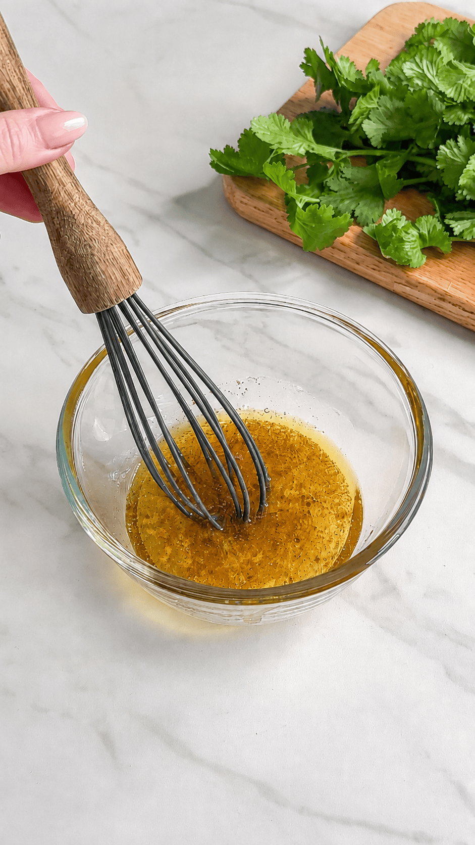 A hand uses a wooden-handled whisk to mix a yellow liquid in a glass bowl, prepping dressing for Mexican Quinoa Salad, with fresh cilantro leaves on a wooden board nearby, all on a white marble surface.