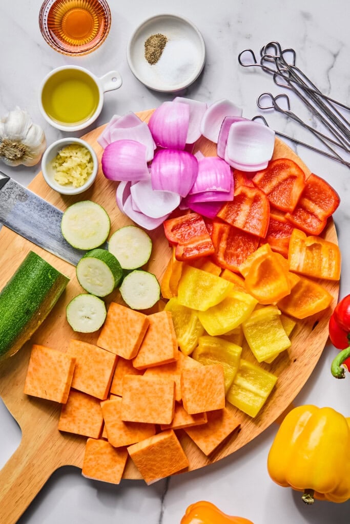 Chopped sweet potato, zucchini, red onion, and red and yellow bell peppers are arranged on a wooden board with a knife, garlic, olive oil, seasoning, and skewers on a marble surface—perfect for assembling vibrant Rainbow Veggie Kebabs.