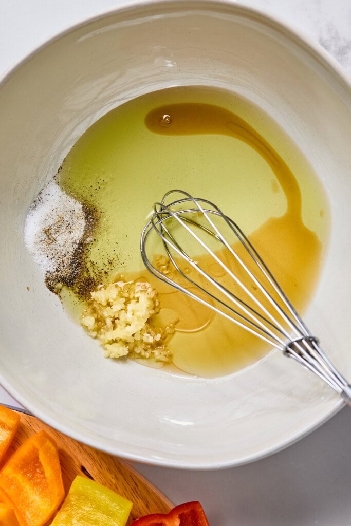 A mixing bowl with olive oil, minced garlic, black pepper, salt, and honey, next to a metal whisk. Chopped yellow and red bell peppers for Rainbow Veggie Kebabs are visible on a wooden board nearby.