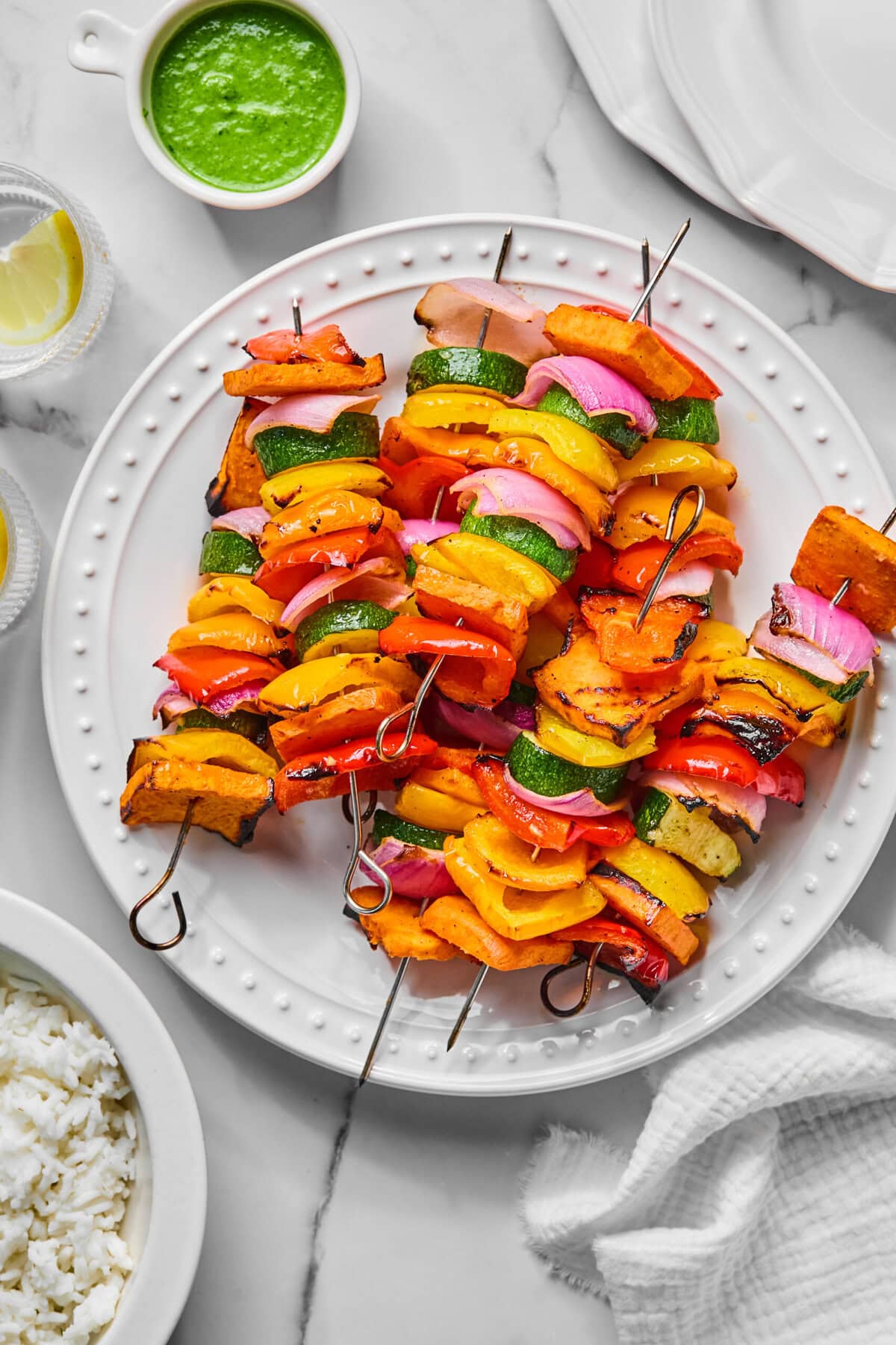 Rainbow Veggie Kebabs featuring grilled zucchini, red onion, and vibrant yellow and red bell peppers are artfully arranged on a white plate, served alongside a flavorful green sauce, fluffy rice, lemon wedges, and a crisp white napkin atop a marble surface.