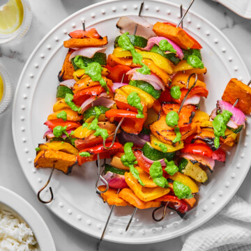 Three Rainbow Veggie Kebabs with grilled vegetables and green sauce on a white plate, surrounded by a bowl of rice, lemon wedges, a cup of green sauce, and stacked empty plates on a marble surface.