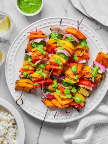 Three Rainbow Veggie Kebabs with grilled vegetables and green sauce on a white plate, surrounded by a bowl of rice, lemon wedges, a cup of green sauce, and stacked empty plates on a marble surface.