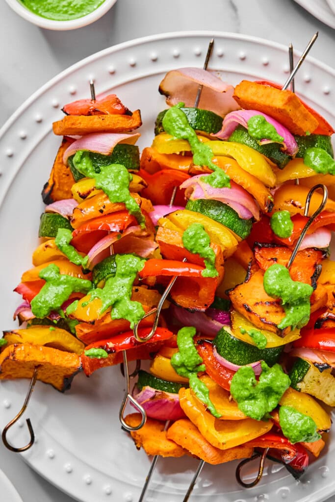 Rainbow Veggie Kebabs featuring colorful skewers of zucchini, bell peppers, onions, and grilled squash are drizzled with vibrant green sauce and served on a white plate.