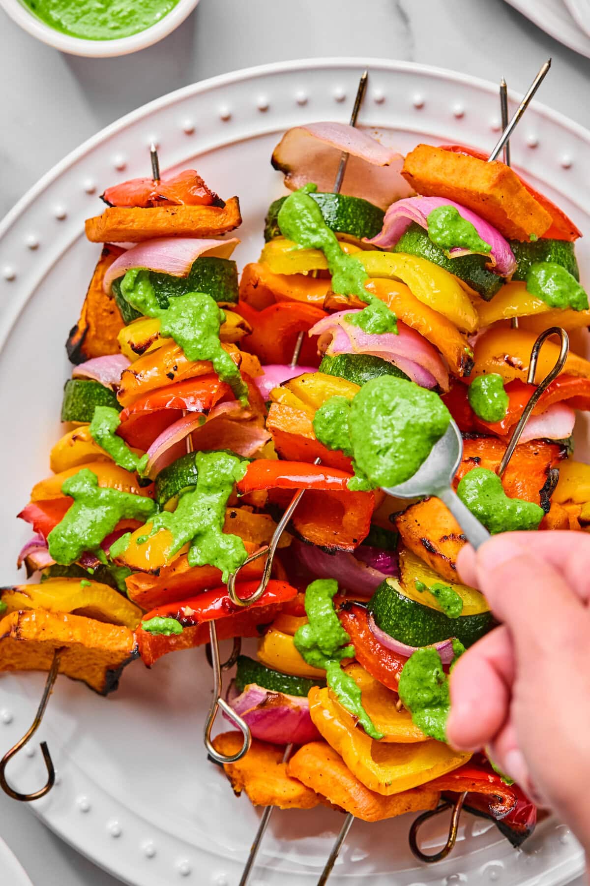 Colorful grilled vegetable skewers with bell peppers, zucchini, onion, and squash are arranged on a white plate. A hand is spooning vibrant jalapeno chimichurri sauce over the skewers. In the background, a small bowl of extra green sauce awaits for drizzling.