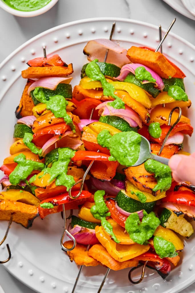 Rainbow Veggie Kebabs featuring colorful skewers with zucchini, bell peppers, onions, and sweet potatoes, drizzled with a bright green sauce and served on a white plate. A hand is spooning more sauce onto the vibrant kebabs.