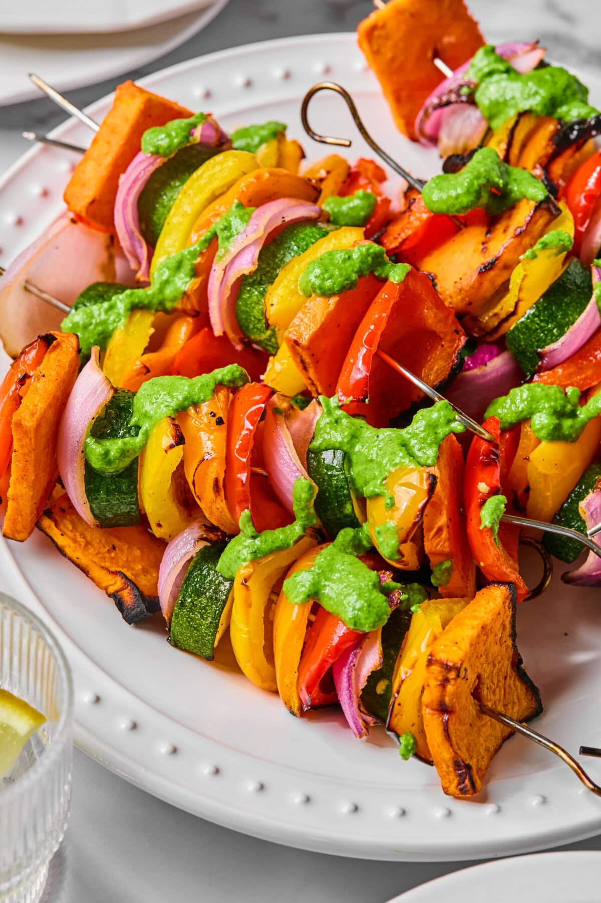 Rainbow Veggie Kebabs featuring grilled skewers with sweet potatoes, bell peppers, zucchini, and red onions, drizzled with green sauce and served on a white plate.