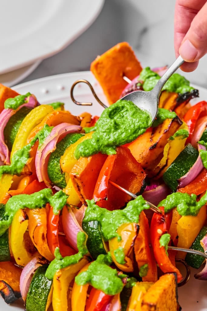 A hand drizzles green sauce over Rainbow Veggie Kebabs—grilled skewers with zucchini, red onion, bell peppers, and squash—artfully arranged on a white plate.