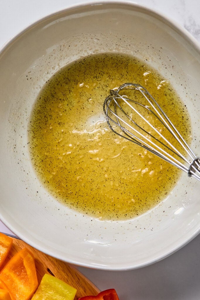 A metal whisk rests in a white bowl containing a yellow, speckled liquid mixture. In the corner, chopped colorful bell peppers are visible on a wooden board, ready to be threaded onto Rainbow Veggie Kebabs.