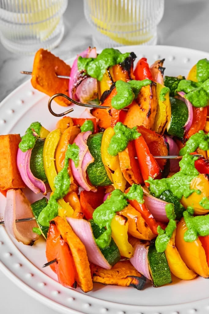 Rainbow Veggie Kebabs are a vibrant medley of red onion, zucchini, bell peppers, and sweet potatoes, all skewered and topped with a fresh green sauce. Served on a white plate with two clear glasses in the background, these colorful kebabs brighten any table.