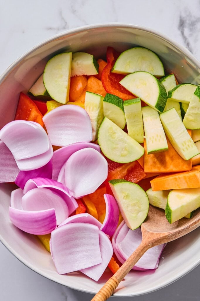 A bowl filled with sliced red onions, cucumbers, carrots, zucchini, and red bell peppers—perfect ingredients for Rainbow Veggie Kebabs—with a wooden spoon resting inside.
