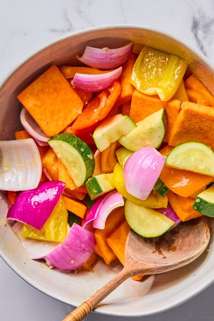 A bowl of chopped vegetables, perfect for Rainbow Veggie Kebabs, including sweet potatoes, zucchini, bell peppers, and red onions, mixed and ready to cook, with a wooden spoon resting on the side.