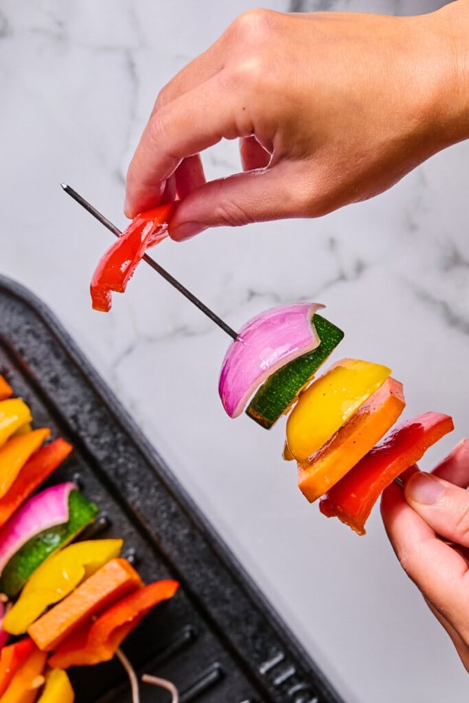A hand assembling a Rainbow Veggie Kebabs skewer with red pepper, onion, zucchini, and yellow pepper over a grill pan with more skewers.