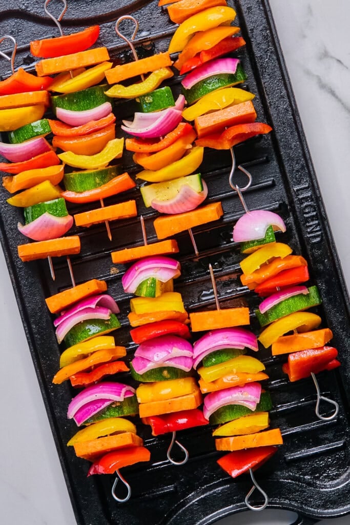 Rainbow Veggie Kebabs featuring colorful vegetable skewers with bell peppers, red onions, and zucchini are artfully arranged on a black grill pan, ready for cooking. The vegetables are cut into chunks and alternated on metal skewers, creating a vibrant feast for the eyes and palate.