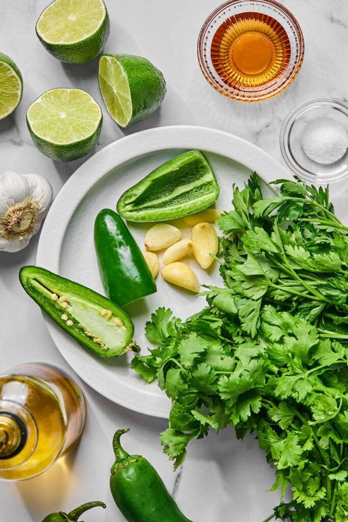A plate with cilantro, garlic cloves, and sliced jalapeño peppers—ingredients that come together perfectly for a fresh jalapeno chimichurri sauce. Surrounding the plate are halved limes, a head of garlic, a bowl of salt, a bowl of amber liquid, and a bottle of oil.
