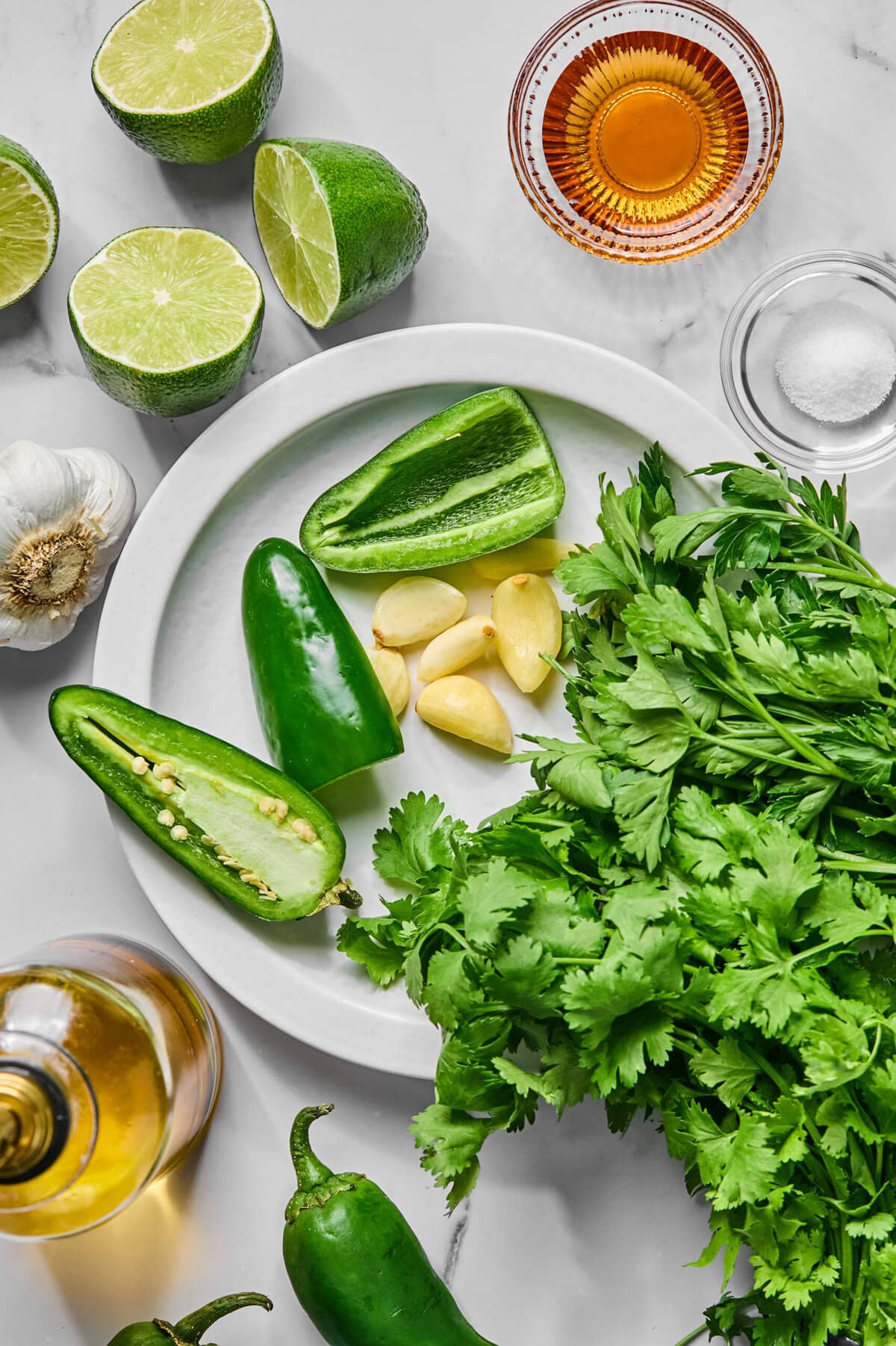 A plate with cilantro, garlic cloves, and sliced jalapeño peppers-ingredients that come together perfectly for a fresh jalapeno chimichurri sauce. Surrounding the plate are halved limes, a head of garlic, a bowl of salt, a bowl of amber liquid, and a bottle of oil.