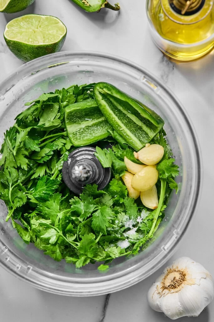 Food processor bowl with cilantro, parsley, sliced jalapeño, garlic cloves, and salt—ingredients ready to be blended into a vibrant jalapeno chimichurri sauce. Nearby are a halved lime, a garlic bulb, and a bottle of olive oil on a white countertop.