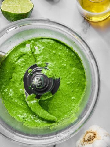 Bright green jalapeno chimichurri sauce in a food processor bowl, surrounded by a halved lime, a bottle of oil, and a head of garlic on a white marble surface.