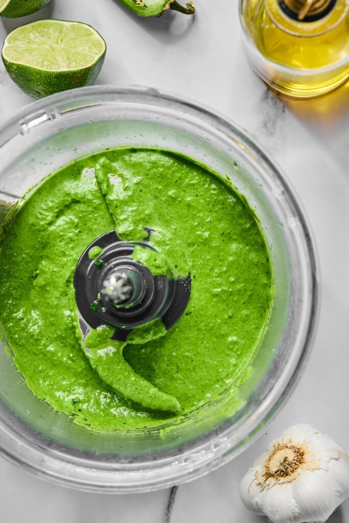 Bright green jalapeno chimichurri sauce in a food processor bowl, surrounded by a halved lime, a bottle of oil, and a head of garlic on a white marble surface.