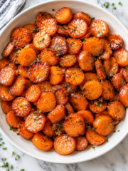 A white bowl filled with maple glazed carrots, garnished with herbs, sits on a marble surface. The carrots are golden and slightly caramelized, with a few sprinkles of green herbs on top.