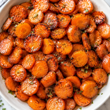A white bowl filled with maple glazed carrots, garnished with herbs, sits on a marble surface. The carrots are golden and slightly caramelized, with a few sprinkles of green herbs on top.