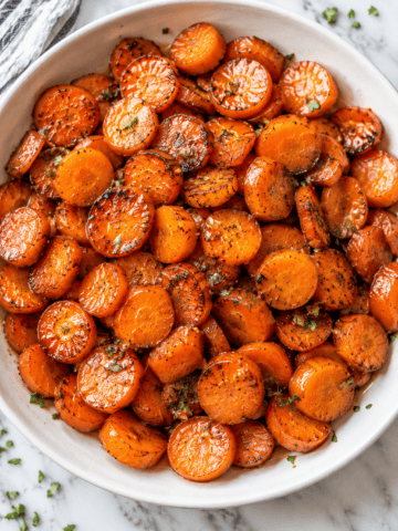 A white bowl filled with maple glazed carrots, garnished with herbs, sits on a marble surface. The carrots are golden and slightly caramelized, with a few sprinkles of green herbs on top.