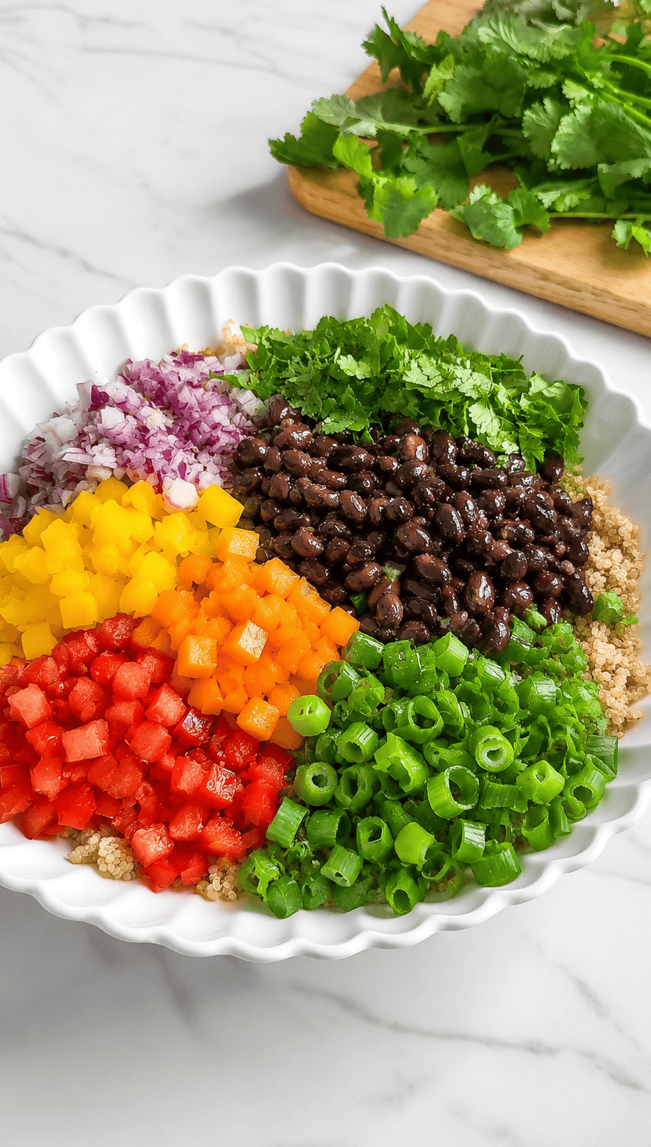 A white scalloped bowl contains vibrant ingredients for a Mexican Quinoa Salad: chopped red onion, yellow and red bell peppers, black beans, green onions, cilantro, and quinoa, with fresh cilantro on a cutting board in the background.