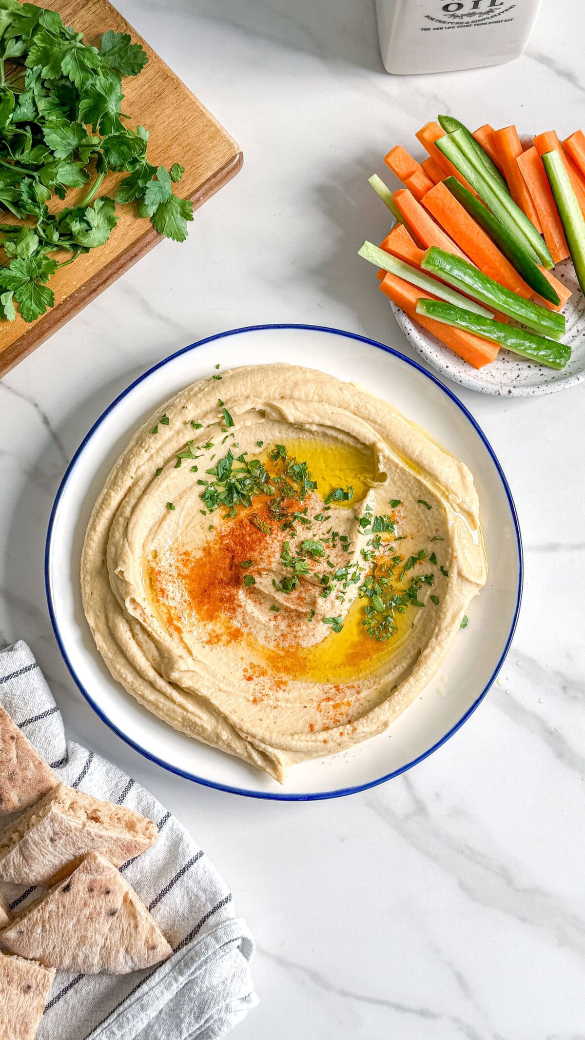 A bowl of easy creamy hummus topped with olive oil, chopped herbs, and paprika sits on a white marble surface. Nearby are pita bread pieces, fresh parsley, and a plate of carrot and cucumber sticks.