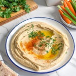 A plate of easy creamy hummus garnished with olive oil, paprika, and herbs sits on a marble surface, surrounded by pita bread, fresh vegetables, herbs on a cutting board, and a ceramic oil bottle.