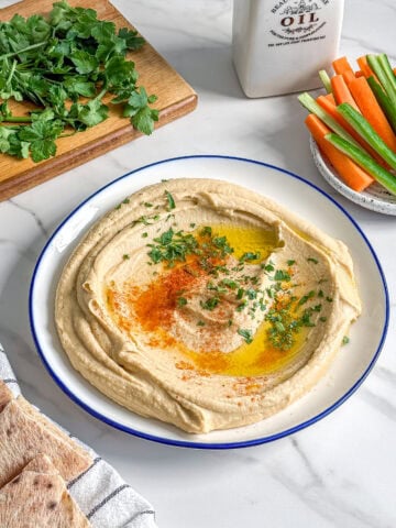 A plate of easy creamy hummus garnished with olive oil, paprika, and herbs sits on a marble surface, surrounded by pita bread, fresh vegetables, herbs on a cutting board, and a ceramic oil bottle.